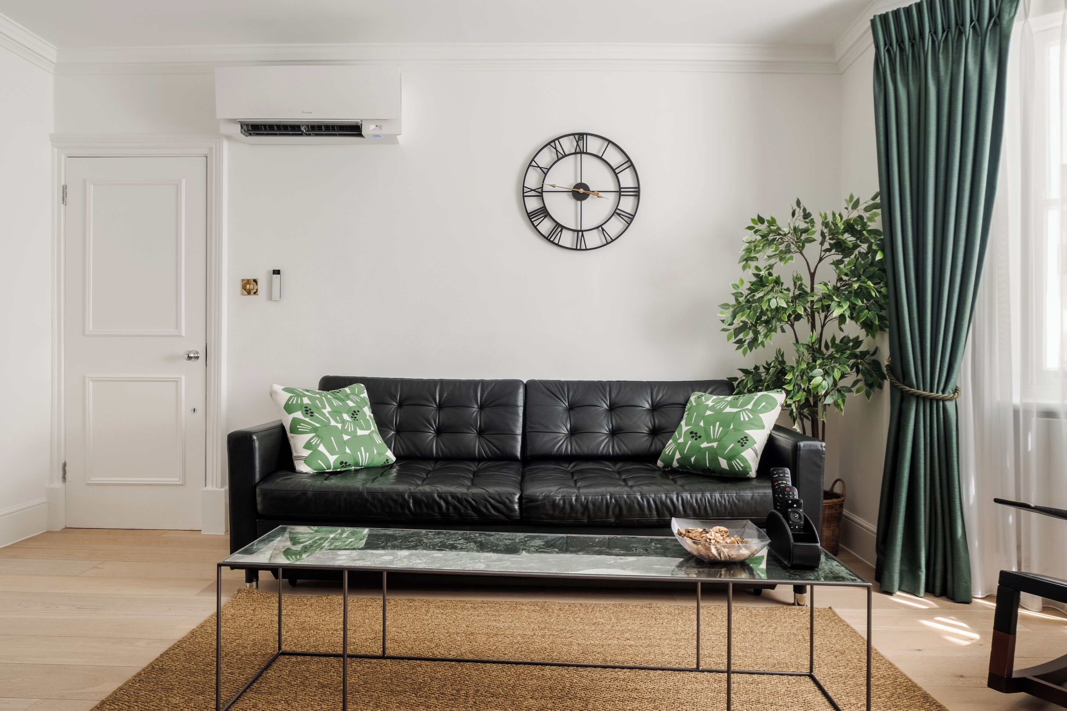 Una sala de estar con un sofá de cuero negro, una mesa de centro de vidrio, un reloj de pared, una planta en maceta y una cortina verde.