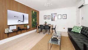 A modern living room with a flat-screen TV, a dining area, a black leather sofa, and a wooden accent wall. - LONDONERS│Emperor's Lounge  (London)