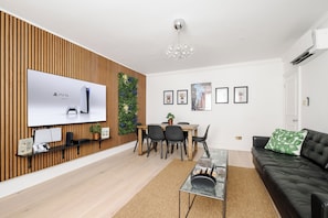 A modern living room with a flat-screen TV, a dining area, a black leather sofa, and a wooden accent wall.