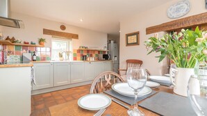 A dining area with a wooden table set for two, a kitchen in the background, and a window with a view of greenery.