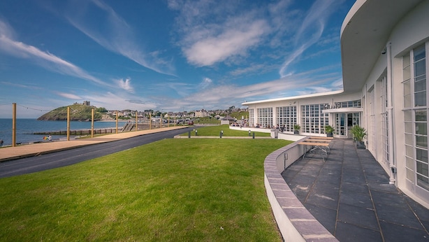Un edificio costero con paseo pavimentado, zonas verdes y vista al mar y colinas distantes.