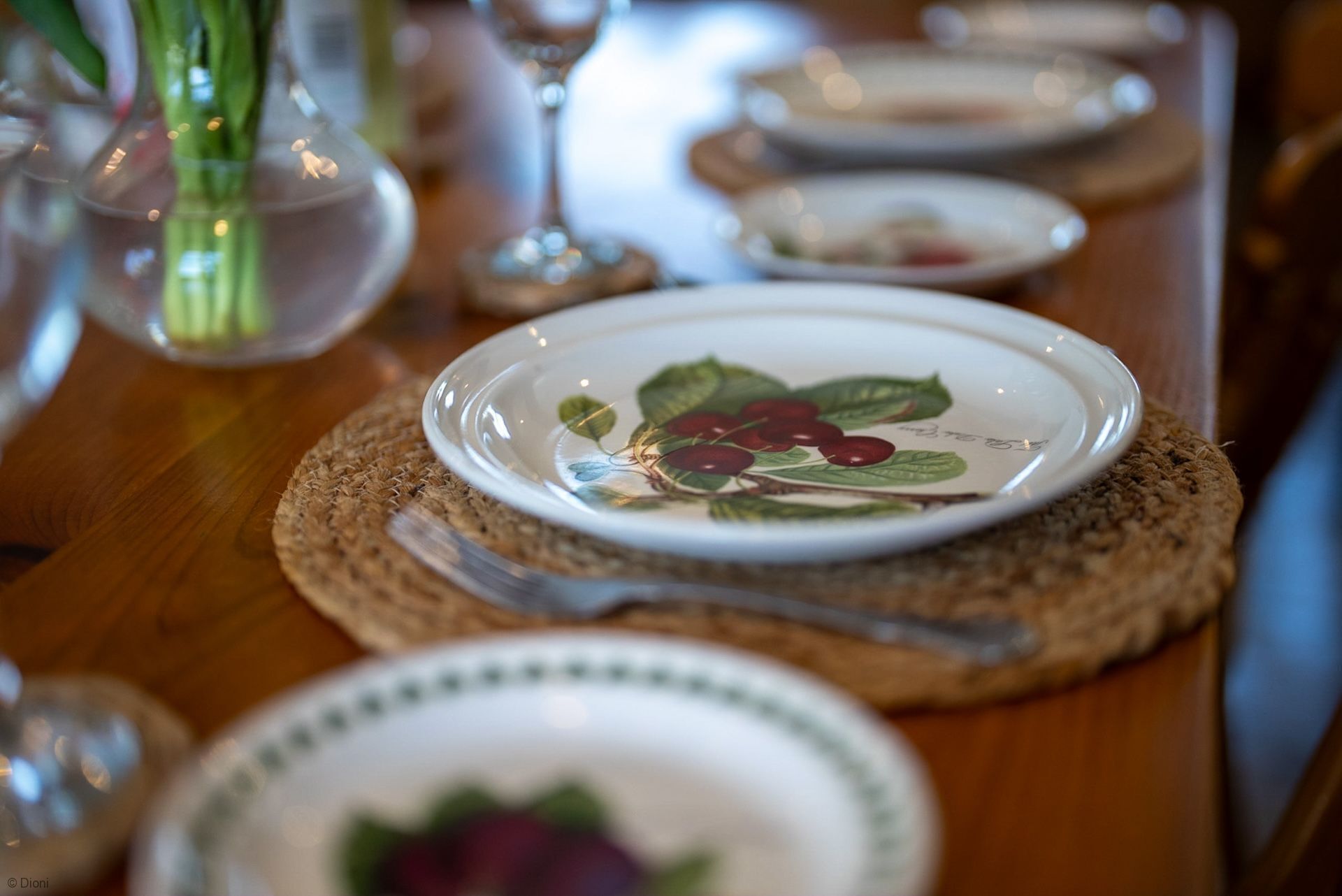 A dining table with plates, cutlery, and a glass vase with green stems.