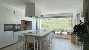 Une cuisine moderne dotée d’un îlot central, de meubles blancs et offrant une vue sur la verdure environnante.