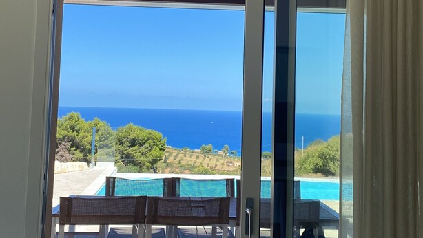 Vue depuis une chambre dotée d’une porte-fenêtre coulissante donnant sur un espace extérieur avec une piscine et une table entourée de chaises.