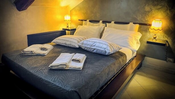 A neatly made bed with pillows and folded towels, bedside lamps, and a dark headboard in a room with a sloped ceiling. - Royal Palace Suite in Piazza Castello (Torino)