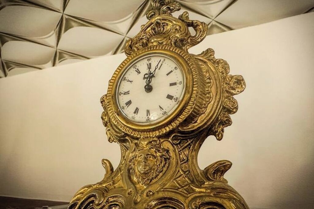Un orologio dorato e decorato con quadrante bianco e numeri romani, posto su una parete spoglia.