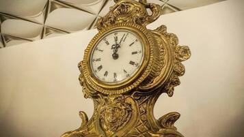 Un orologio dorato e decorato con quadrante bianco e numeri romani, posto su una parete spoglia.