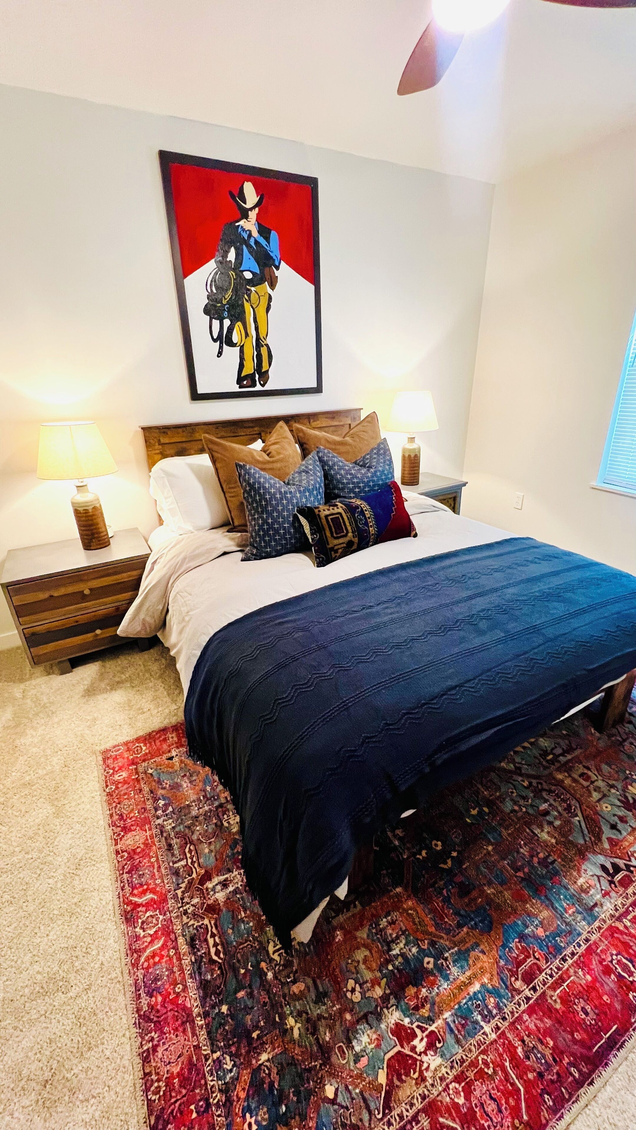 A bedroom with a bed, two bedside tables, a nightstand, a patterned rug, and a wall-mounted artwork.