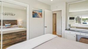 A bedroom with a wooden headboard bed, a bedside table with a lamp, a painting on the wall, and a bathroom with a mirror and sink. - Beautiful 2 Bed Beach Condo #104 2 Bedroom Condo by RedAwning (Carpinteria)