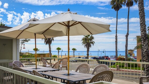 An outdoor seating area with tables and chairs, a large umbrella, and a view of the beach and ocean. - Beautiful 1 Bed Beach Condo #122 1 Bedroom Condo by RedAwning (Carpinteria)