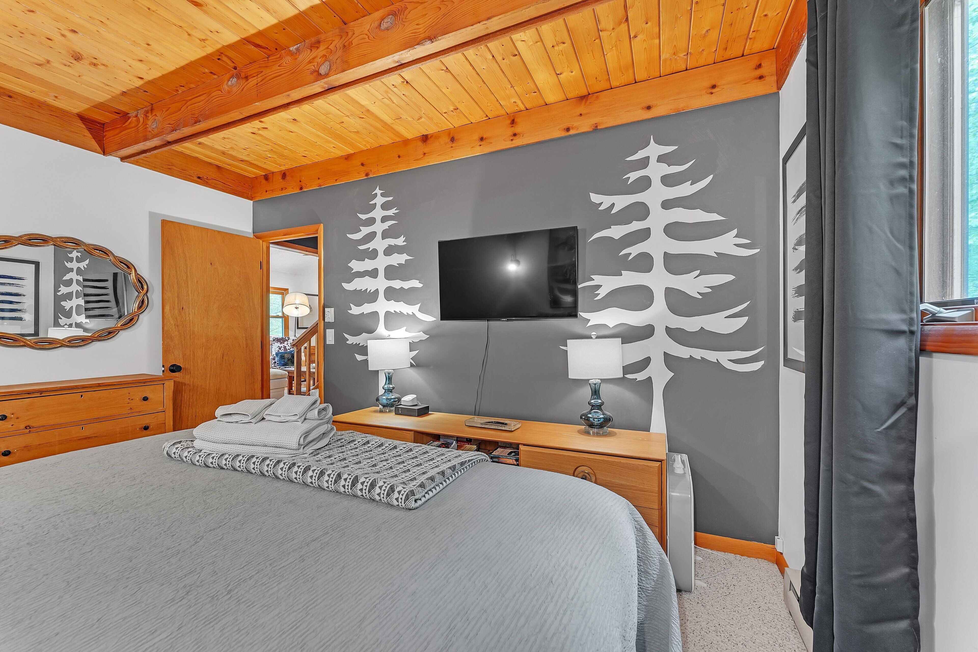 A bedroom with a wooden ceiling, a bed with a grey blanket, two bedside tables with lamps, a TV on the wall, and a mirror on the left.