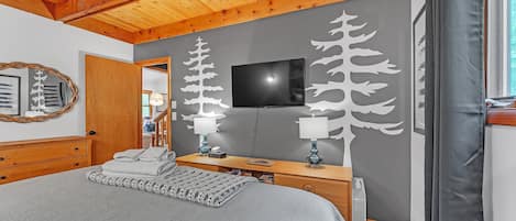 Un dormitorio con techo de madera, una cama con manta gris, dos mesitas de noche con lámparas, un televisor en la pared y un espejo a la izquierda.