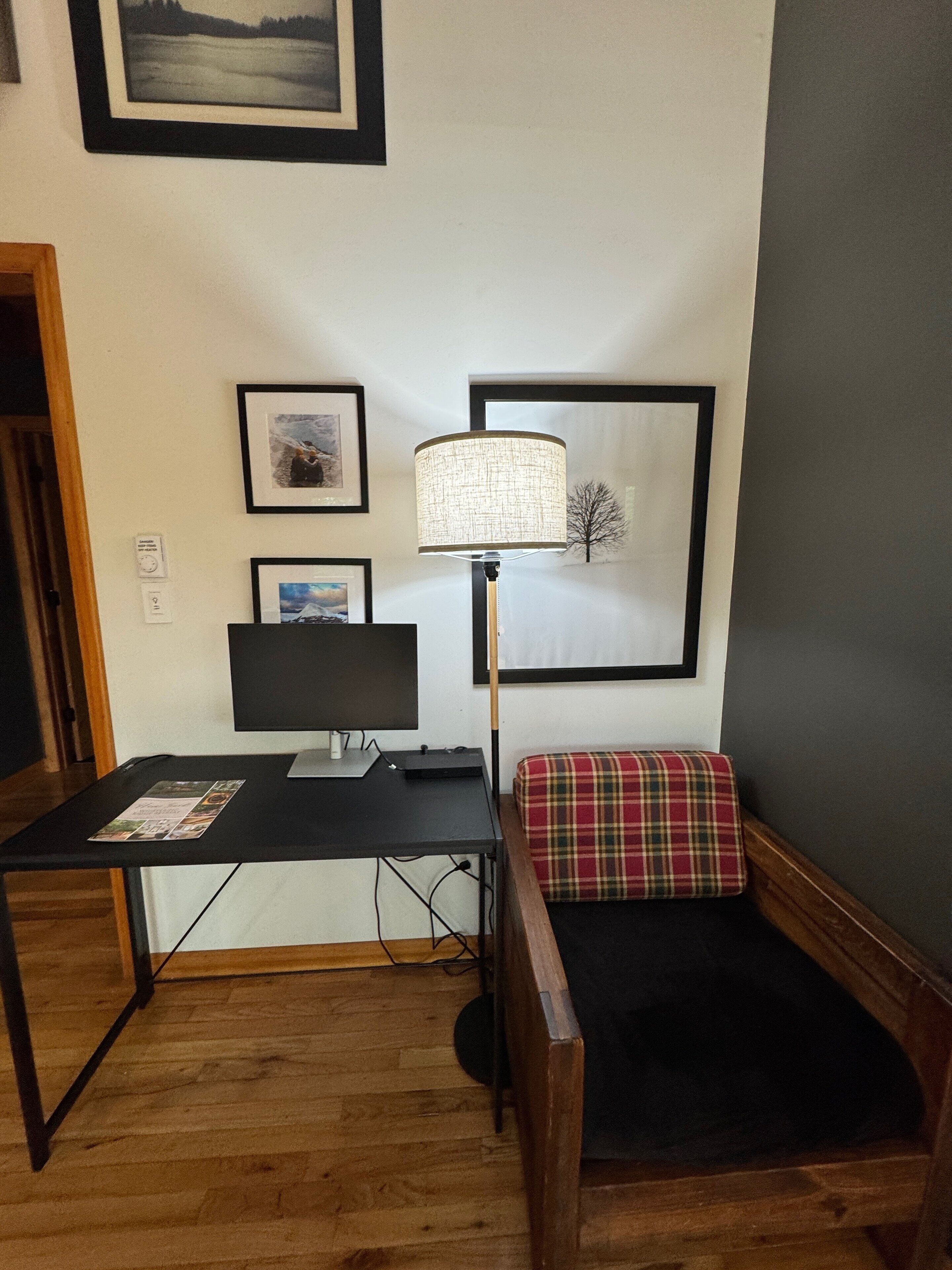 A room with a wooden desk, a black monitor, a lamp, and a plaid cushion on a bench.