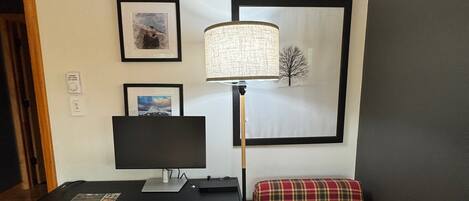 A room with a wooden desk, a black monitor, a lamp, and a plaid cushion on a bench.