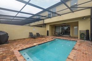 A covered outdoor swimming pool with lounge chairs, a table, and a grill.