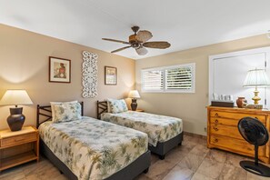 A hotel room with two beds, a ceiling fan, a window with blinds, a wooden dresser, and a nightstand with a lamp.