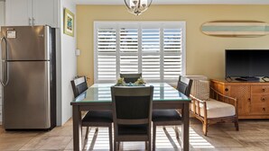 A modern kitchen with a stainless steel refrigerator, a dining table with chairs, a chandelier, and a television on a wooden cabinet. - Kihei Beach, #210 2 Bedroom Condo (Kihei)