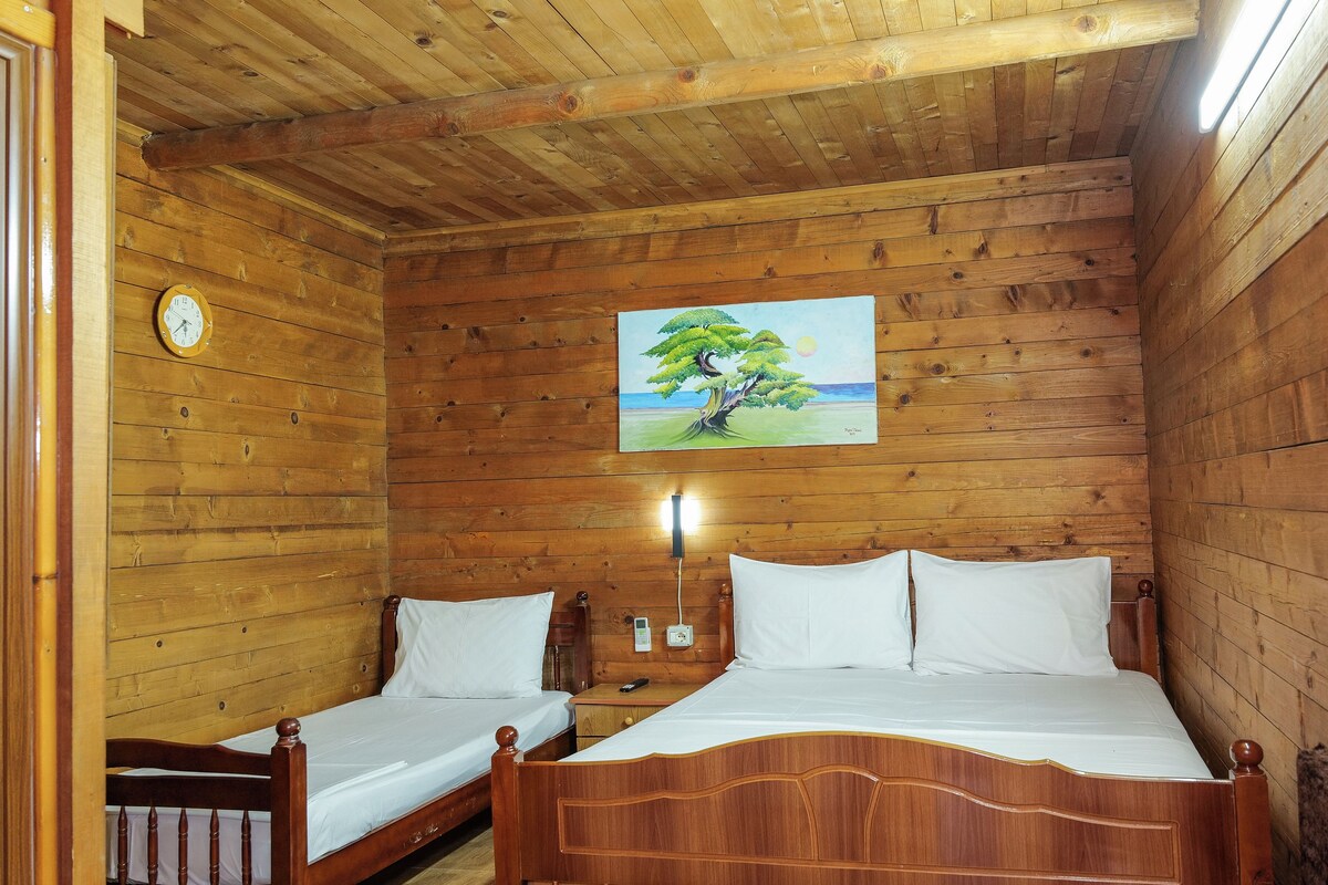 A wooden cabin room with two single beds, a wooden ceiling, a painting of a tree, and a clock on the wall.