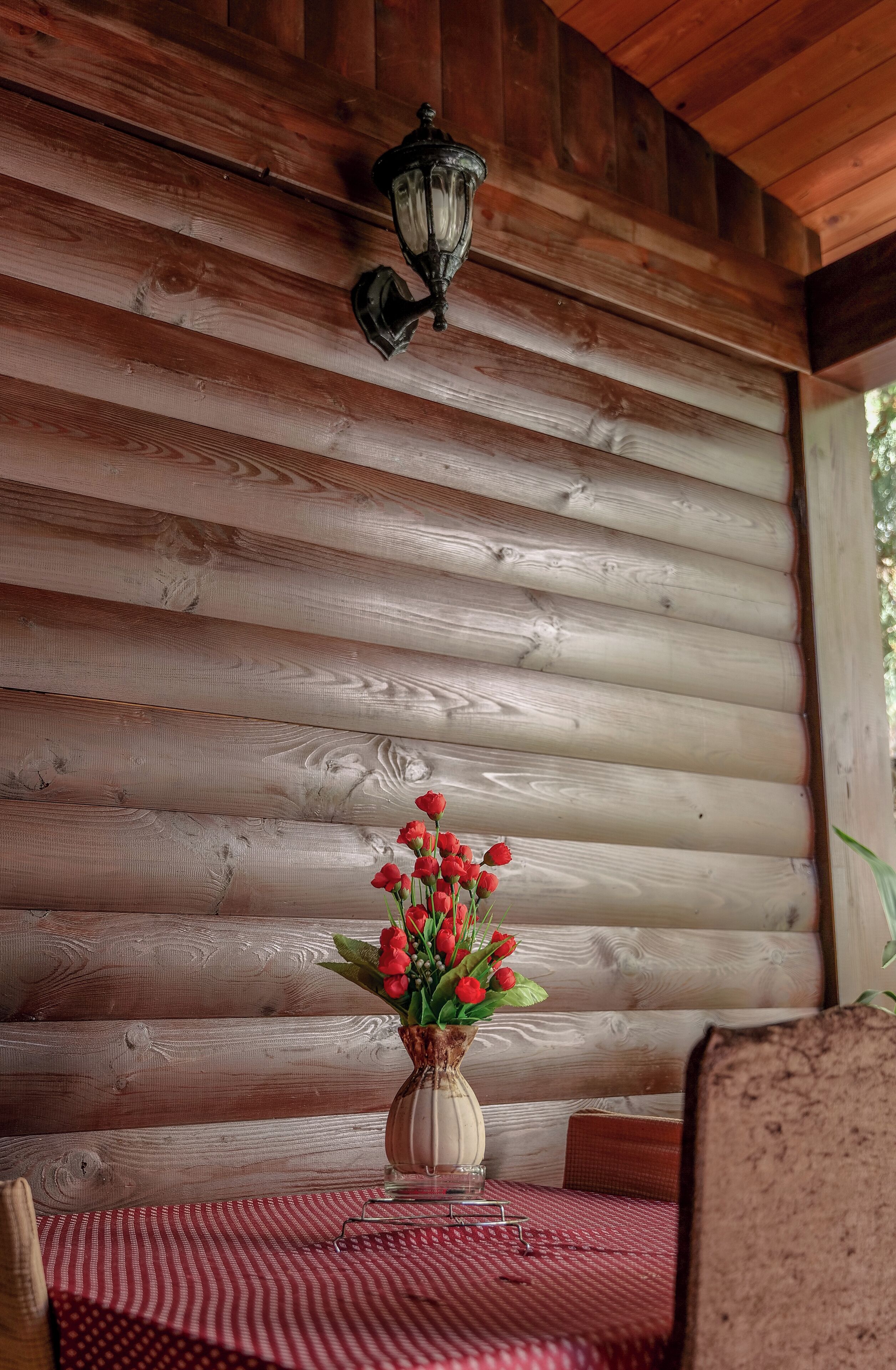 A wooden log cabin interior with a table set for dining, a vase with red flowers, and a wall-mounted lantern.