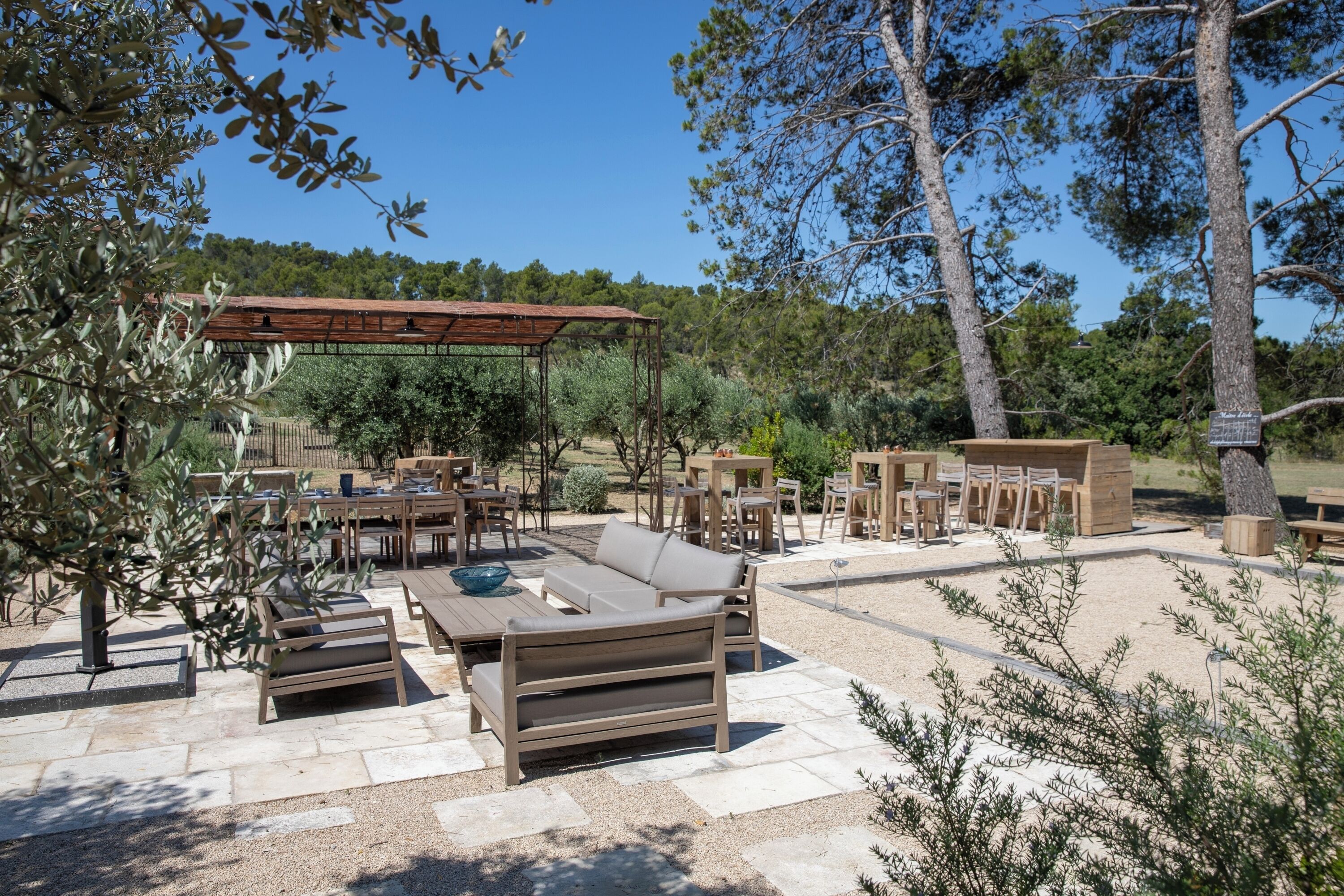 Een terras buiten met houten meubels, waaronder banken en tafels, omringd door olijfbomen en een pergola.