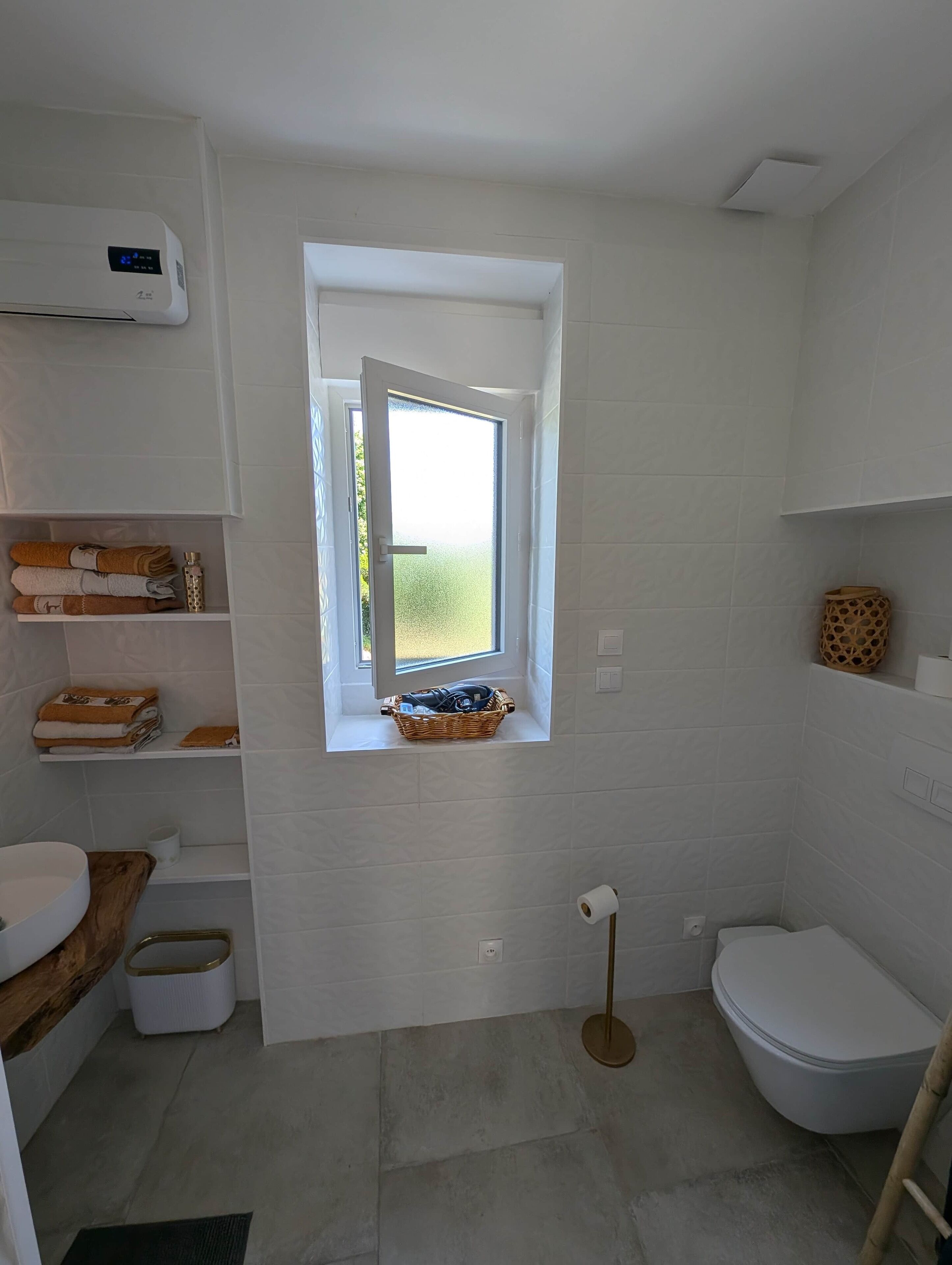 A modern bathroom with a built-in window, a white toilet, a sink with a wooden countertop, and a shelf with towels and toiletries.