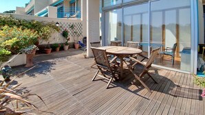Terraza de madera con una mesa y sillas, rodeada de plantas en macetas y una puerta de cristal que da a un balcón.