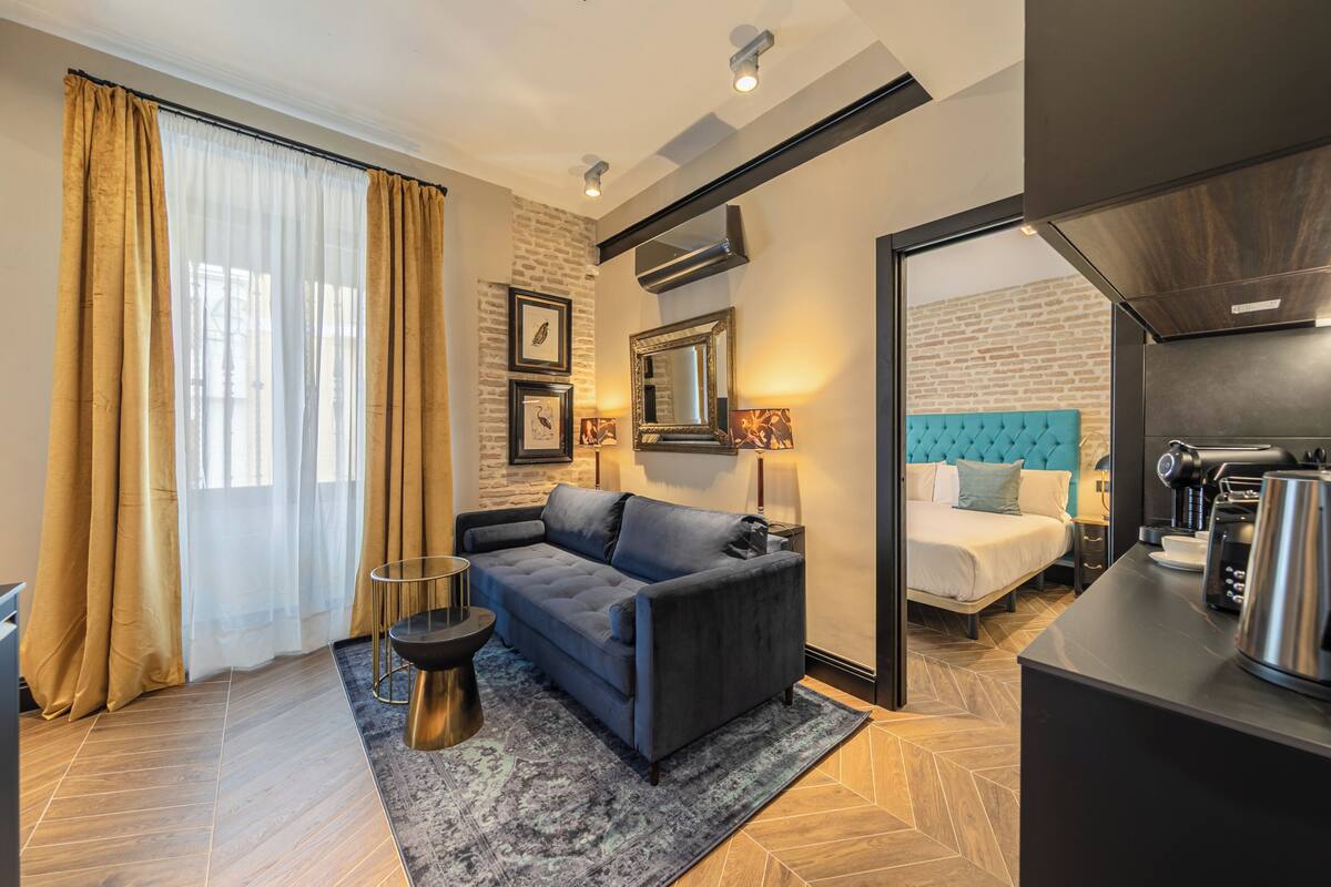 Una habitación de hotel moderna con sofá, cama con cabecera azul, cafetera y chimenea.