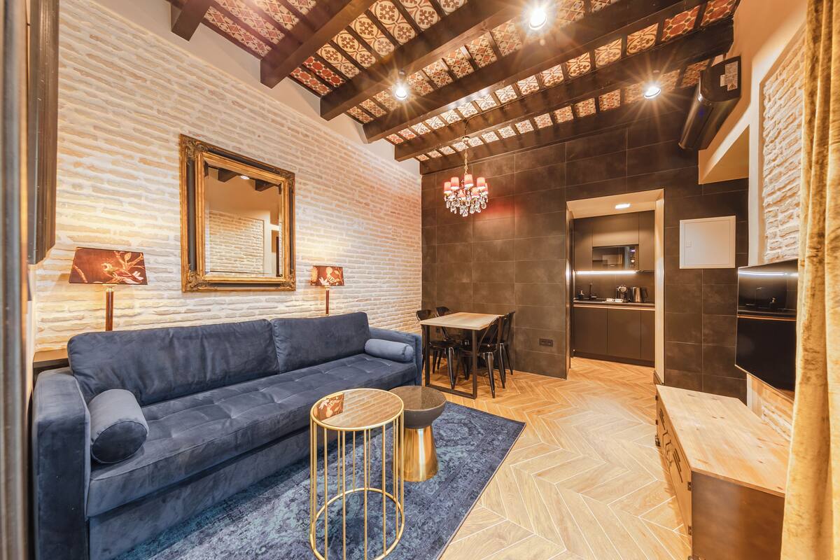 Un salón moderno con un sofá azul oscuro, una mesa de centro redonda y una alfombra estampada. Se observa una zona de comedor con una mesa y sillas de madera, y una cocina con baldosas oscuras y electrodomésticos modernos.