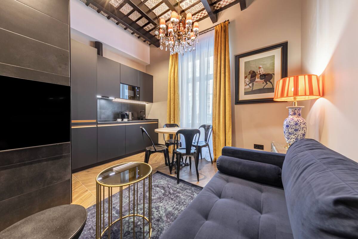 Una sala de estar moderna con un sofá, un televisor, un comedor con sillas y una mesa, y una cocina con armarios oscuros y una araña.