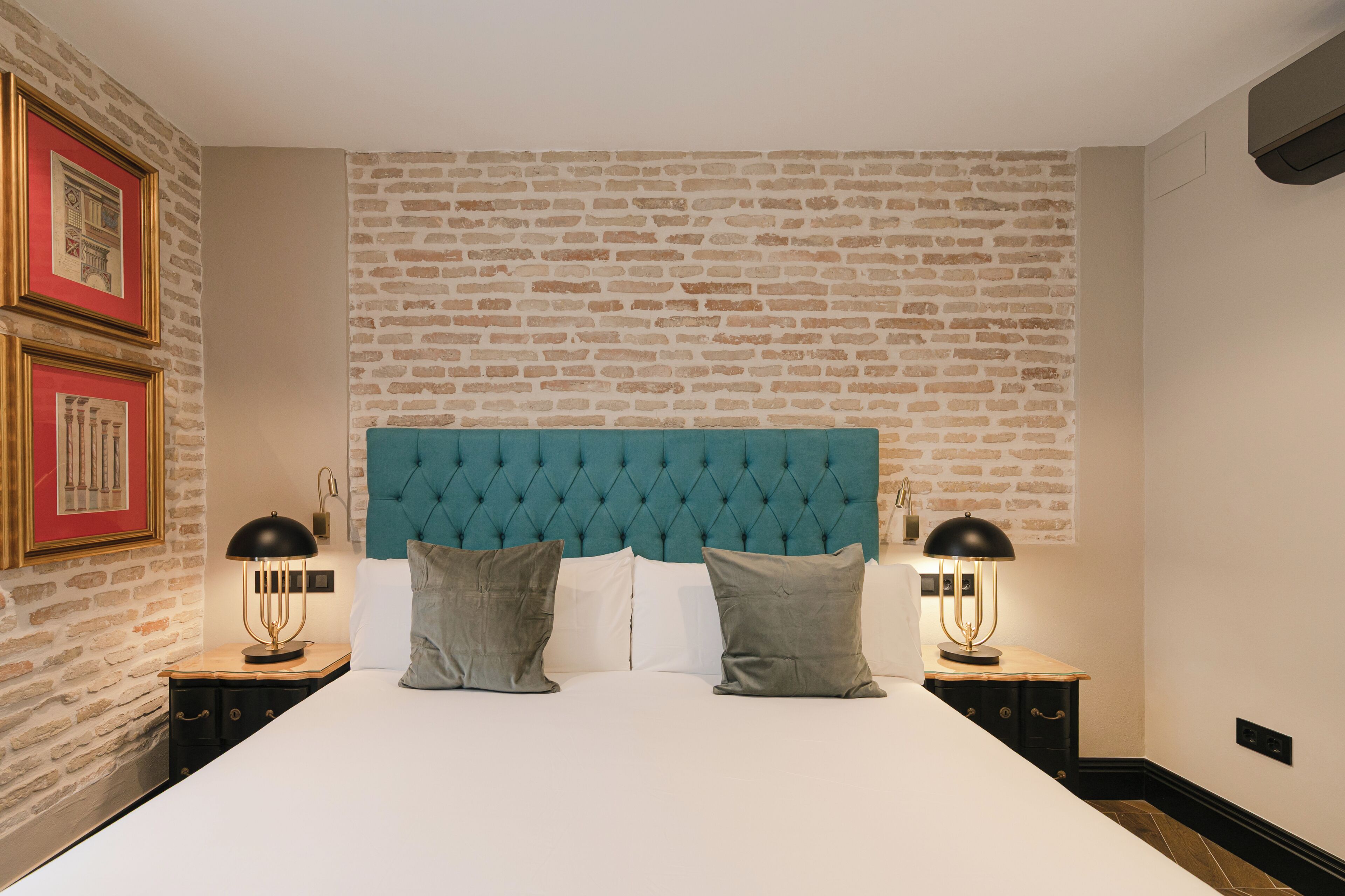 A bedroom with a brick-patterned wall, a tufted teal headboard, two bedside tables with lamps, and a bed with white linens and gray pillows.
