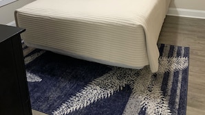 Una cama bien hecha con cabecera, una lámpara de noche y una alfombra estampada sobre un suelo de madera.