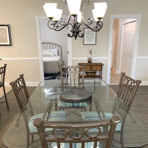 A dining area with a glass-top table, cushioned chairs, a chandelier, and a bed in the background.