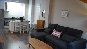 A modern living room with a dark grey sofa, a wooden bench, and a white dining area with chairs. - Apartamentos Altar Suites Conde Luna (León)