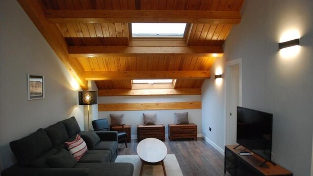A modern living room with a high ceiling, wooden beams, a sofa set, a round coffee table, a TV stand, and a skylight. - Apartamentos Altar Suites Conde Luna (León)