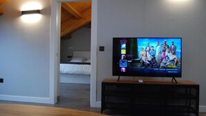 A modern living room with a flat-screen TV on a wooden stand, a white bed in the background, and a wooden ceiling. - Apartamentos Altar Suites Conde Luna (León)