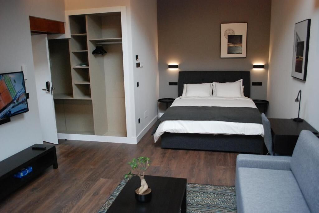 A modern hotel room with a large bed, a grey sofa, a small table with a plant, a wardrobe, and a painting on the wall.