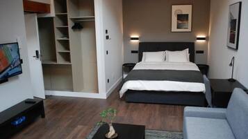 A modern hotel room with a large bed, a grey sofa, a small table with a plant, a wardrobe, and a painting on the wall.