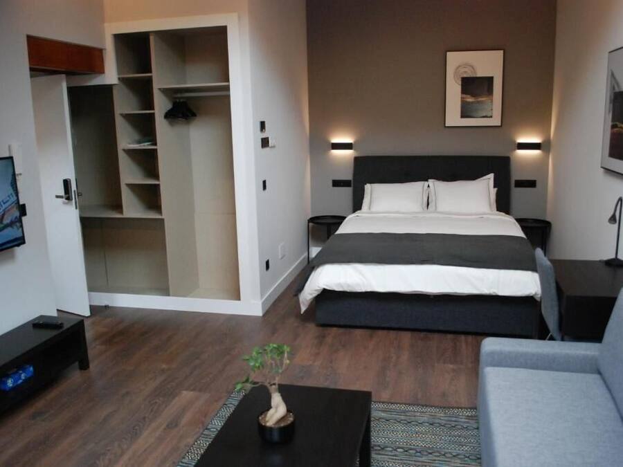 A modern hotel room with a large bed, a grey sofa, a small table with a plant, a wardrobe, and a painting on the wall.