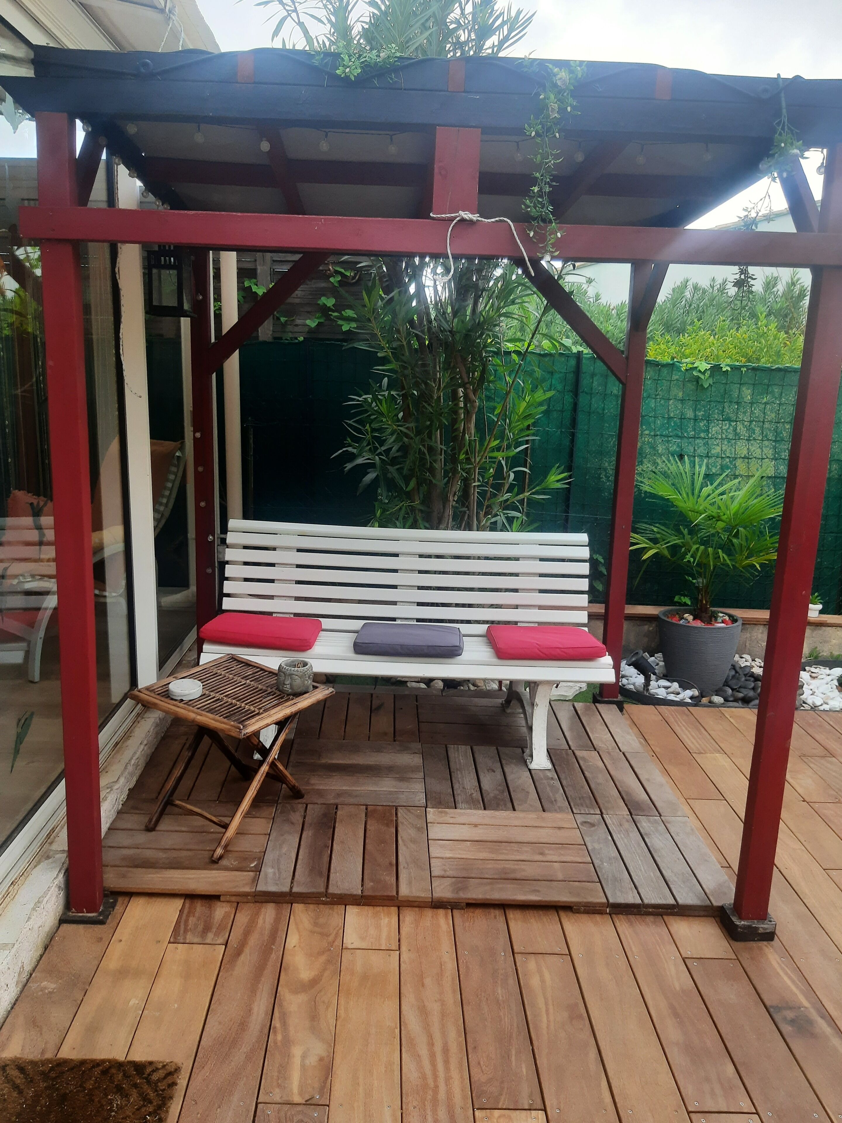 A wooden deck with a white bench under a red pergola, surrounded by potted plants and a green fence.
