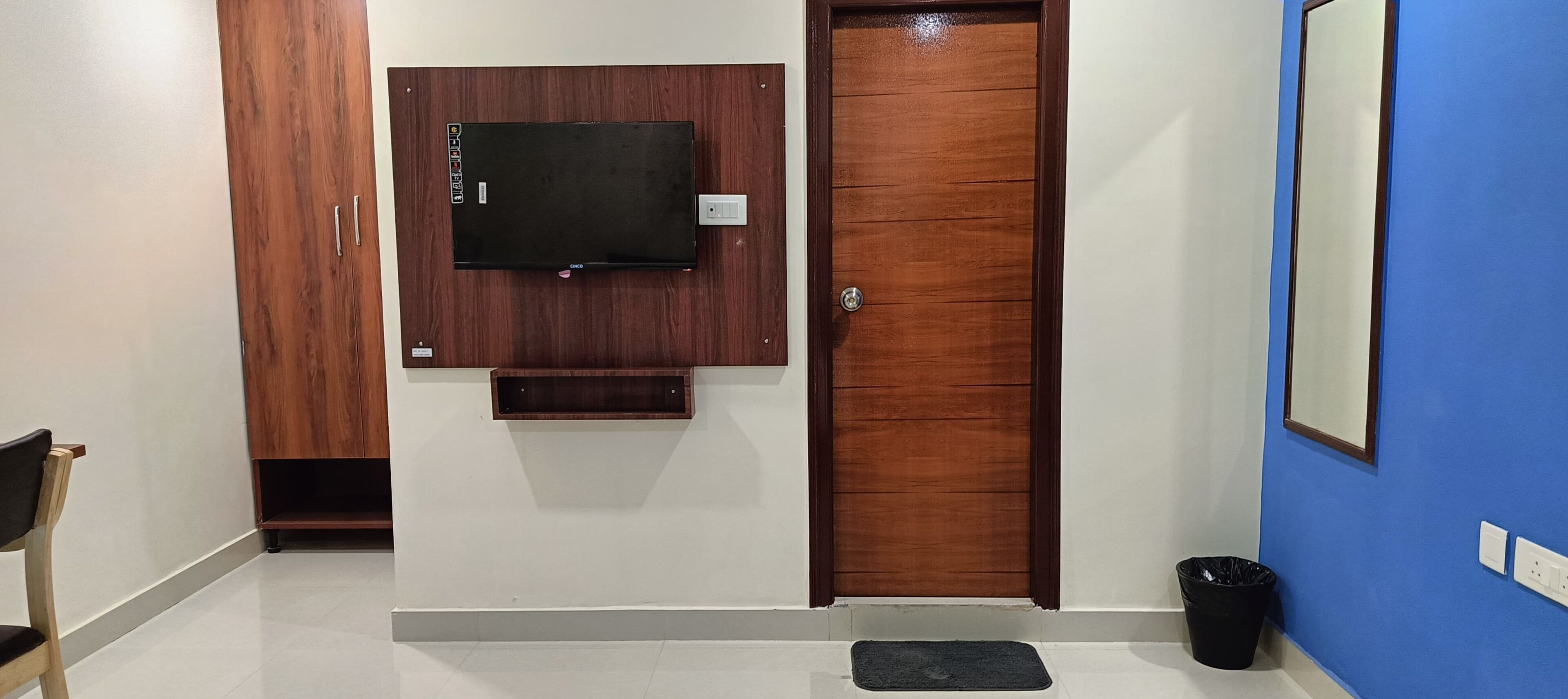 A hallway with a wooden door, a mounted television, a trash bin, and a small shelf.