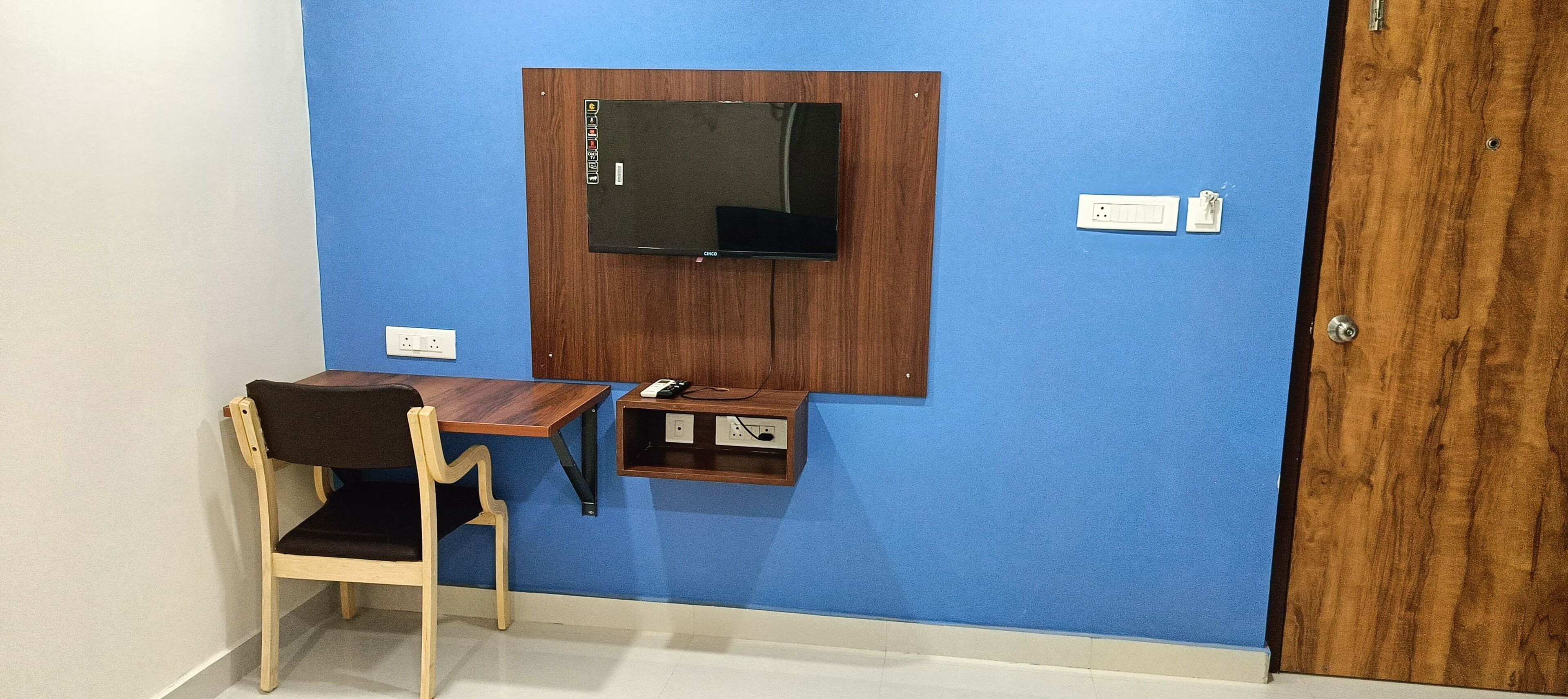 A small room with a blue wall, a wooden desk, a chair, a television mounted on the wall, and a door.