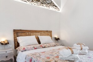A bedroom with a wooden headboard, white bedding, a nightstand with a lamp, and a wall-mounted light fixture.