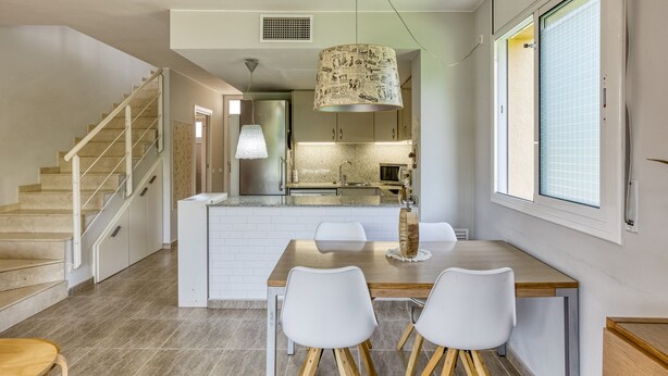 Een moderne keuken met een centraal eiland, witte stoelen, een houten eettafel en een trap.