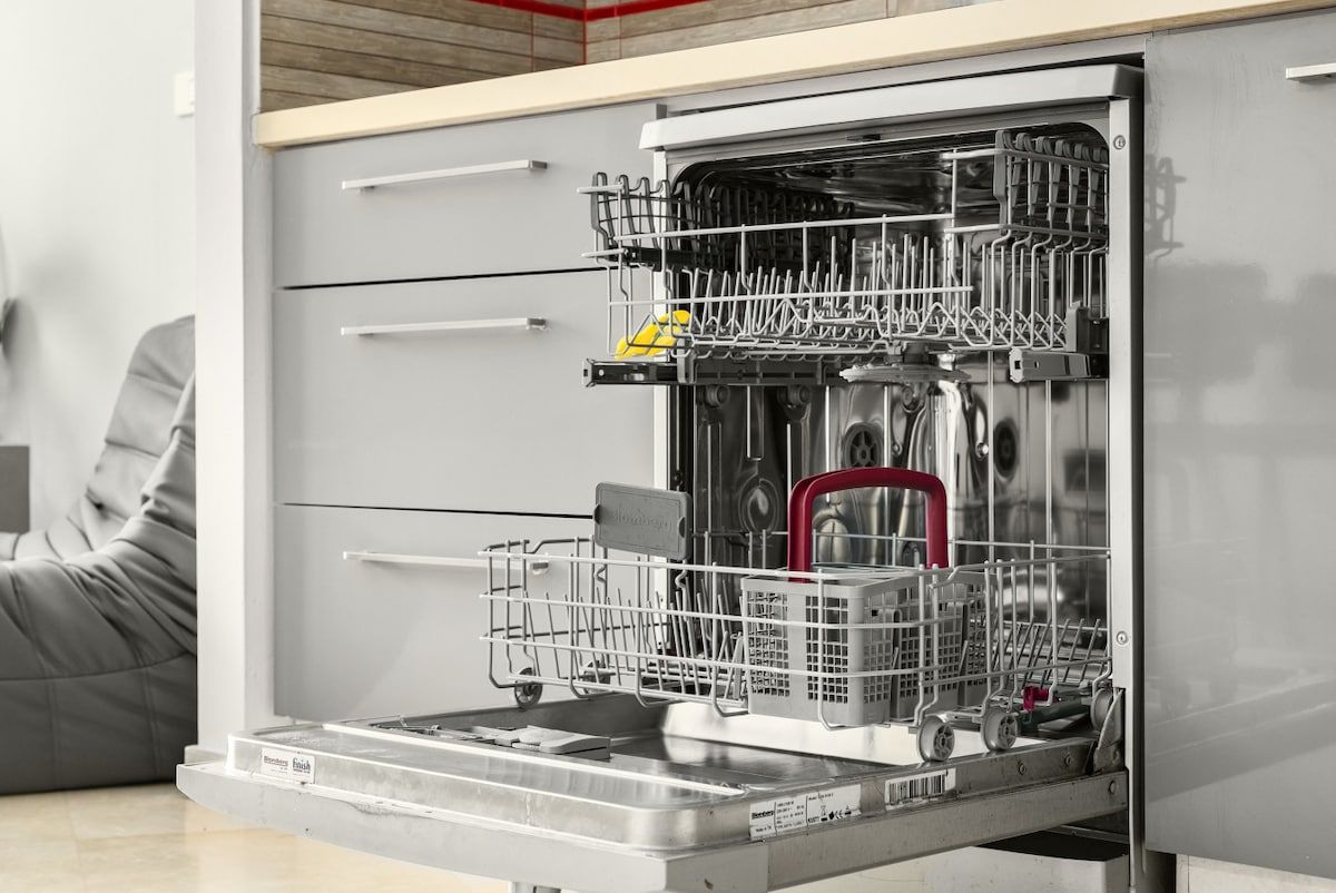 An open dishwasher with racks and utensils inside, a grey cabinet with drawers, and a grey sofa in the background.