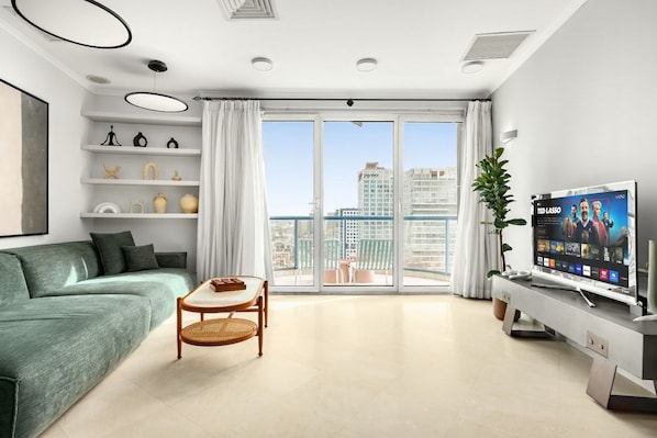 A modern living room with a sofa, a small table, a TV, and a balcony with a view of the city.