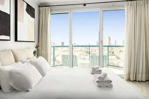 A hotel room with a large bed, two chairs, a balcony with a city view, and a painting on the wall.