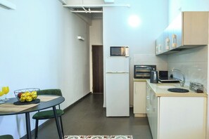 Eine kompakte Küche mit Kühlschrank, Mikrowelle und Backofen. Ein Esstisch mit Stühlen und einer Obstschale sind vorhanden.