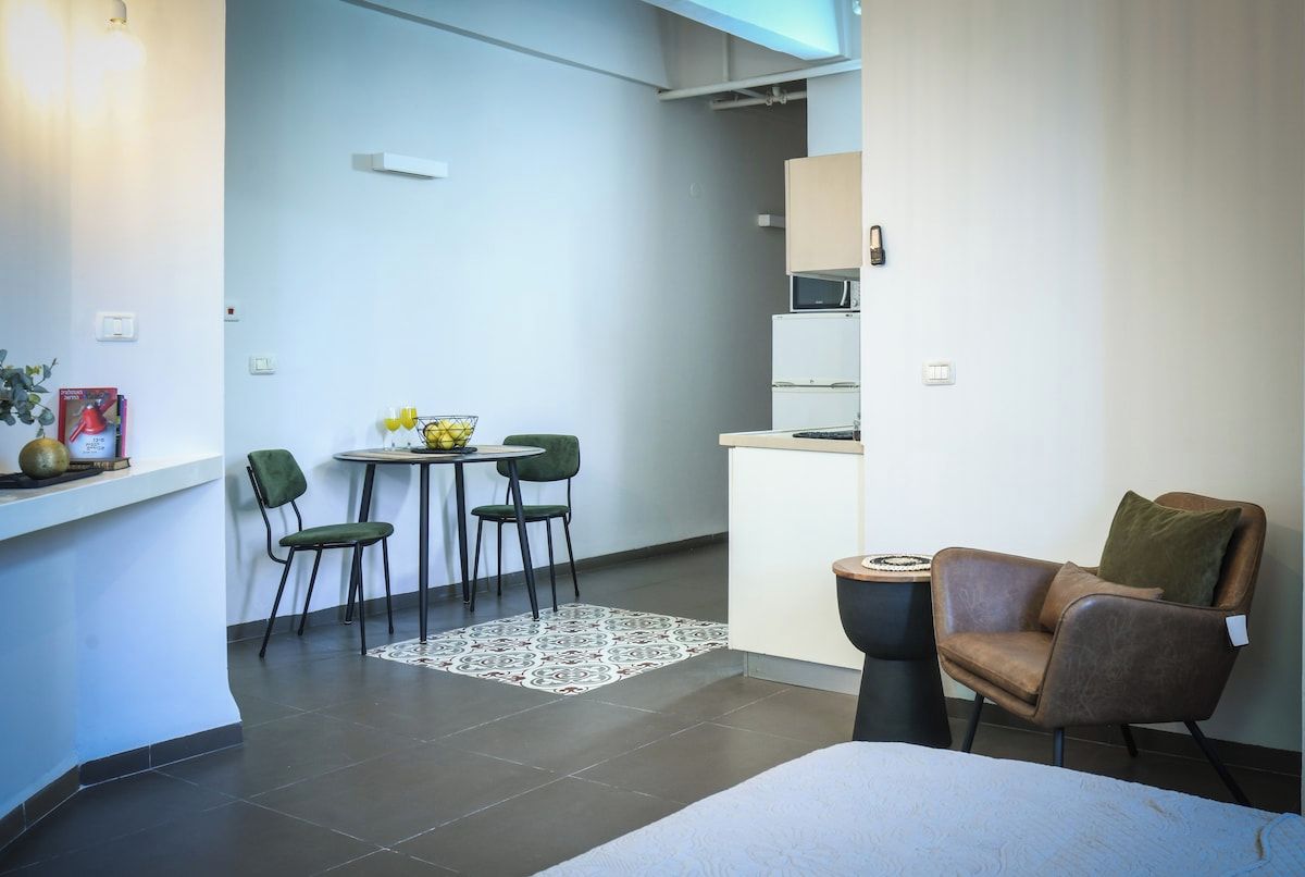 A compact living space with a bed, a dining area with chairs, a small kitchen, and a window.