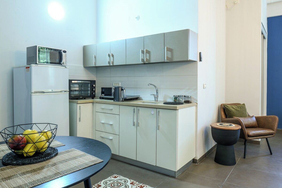 A compact kitchen with a microwave, toaster, and kettle on the countertop, a small dining table with a fruit bowl, and a chair with a cushion.