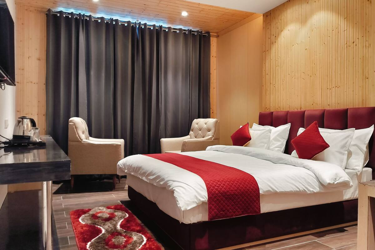 A modern hotel room with a large bed, two armchairs, a wooden headboard, and a flat-screen TV mounted on the wall.